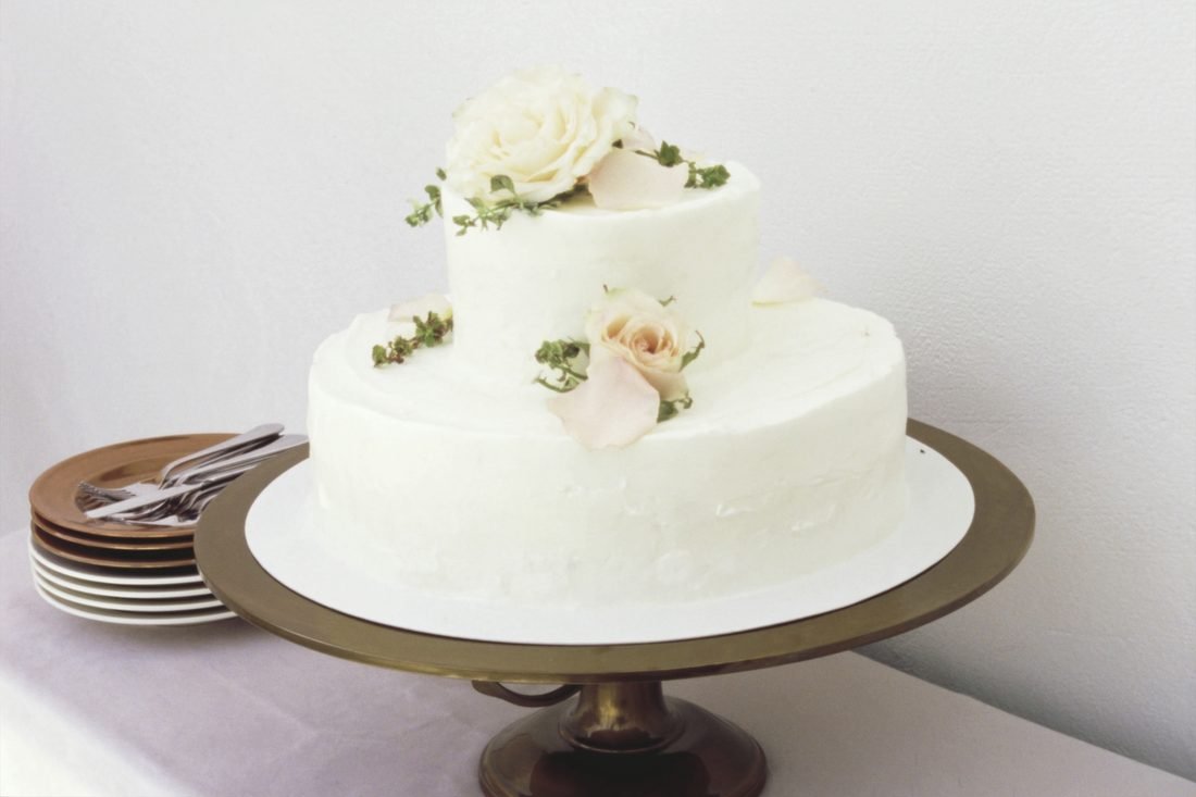 A two-tier iced cake on a brass stand with fresh roses, beside a stack of plates and forks