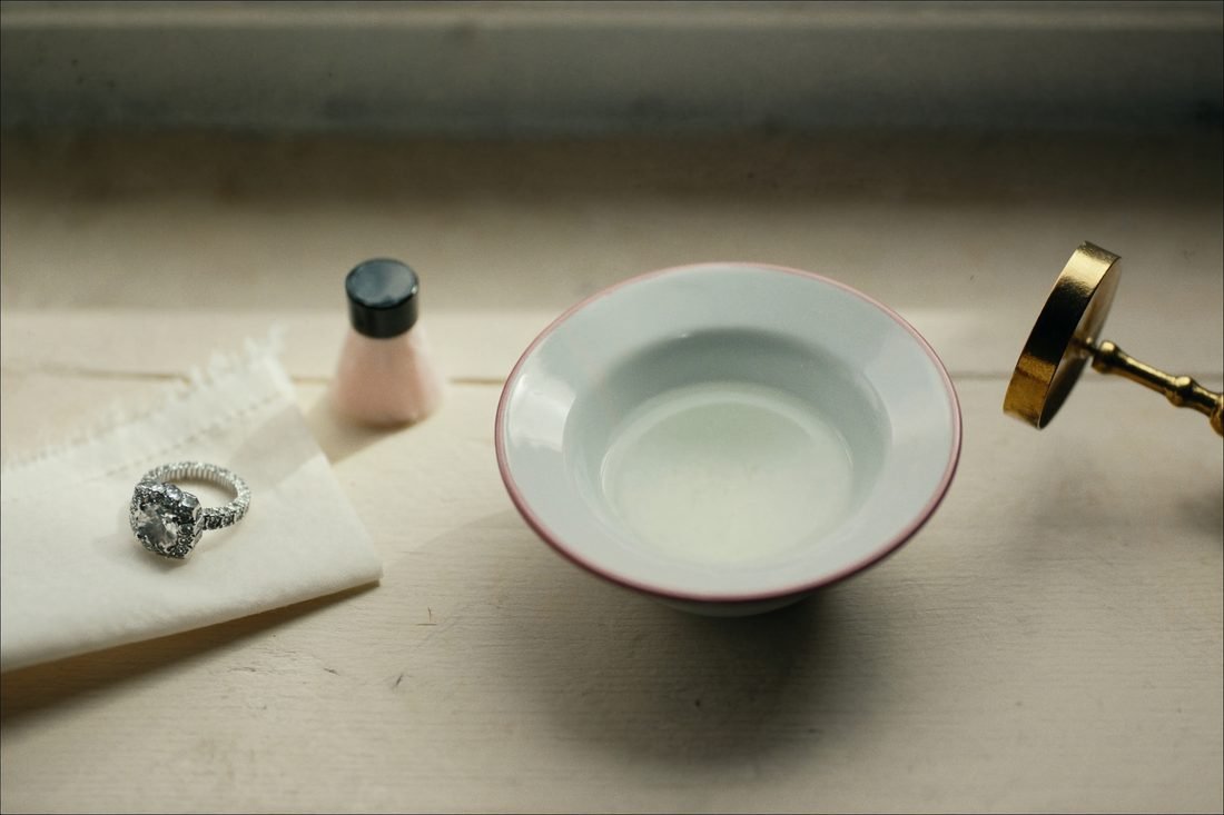 A diamond ring on a polishing cloth beside a soft brush, an enamel bowl and a loupe
