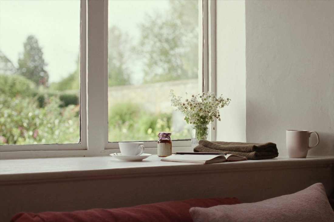 A cottage window seat with a cup of tea, a closed notebook and a jar of hedgerow flowers