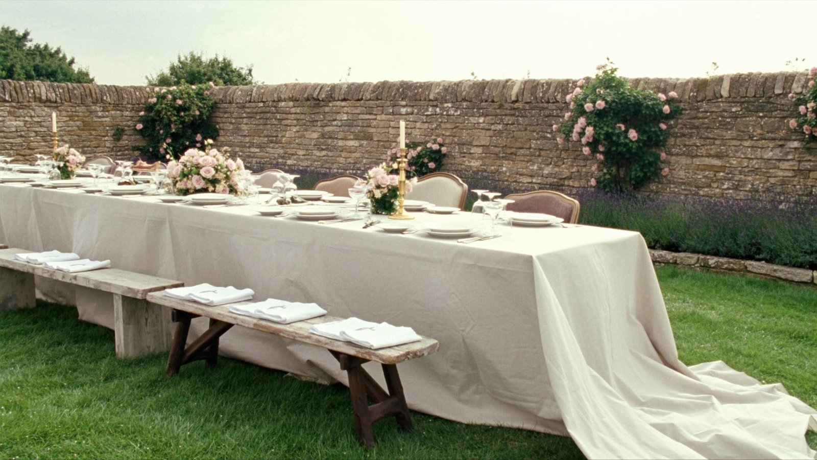 A long table laid for a wedding breakfast in a walled Cotswold garden, climbing roses on the stone and lavender along the border