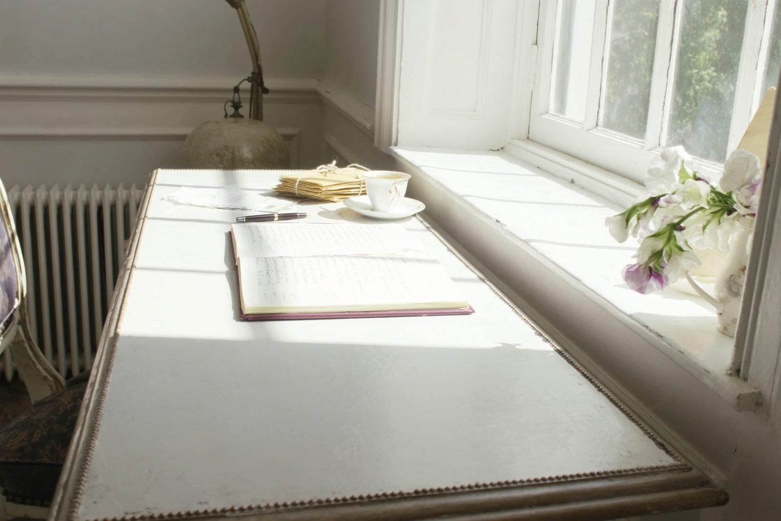 A pale desk under a sunny window with an open ledger, a fountain pen and envelopes