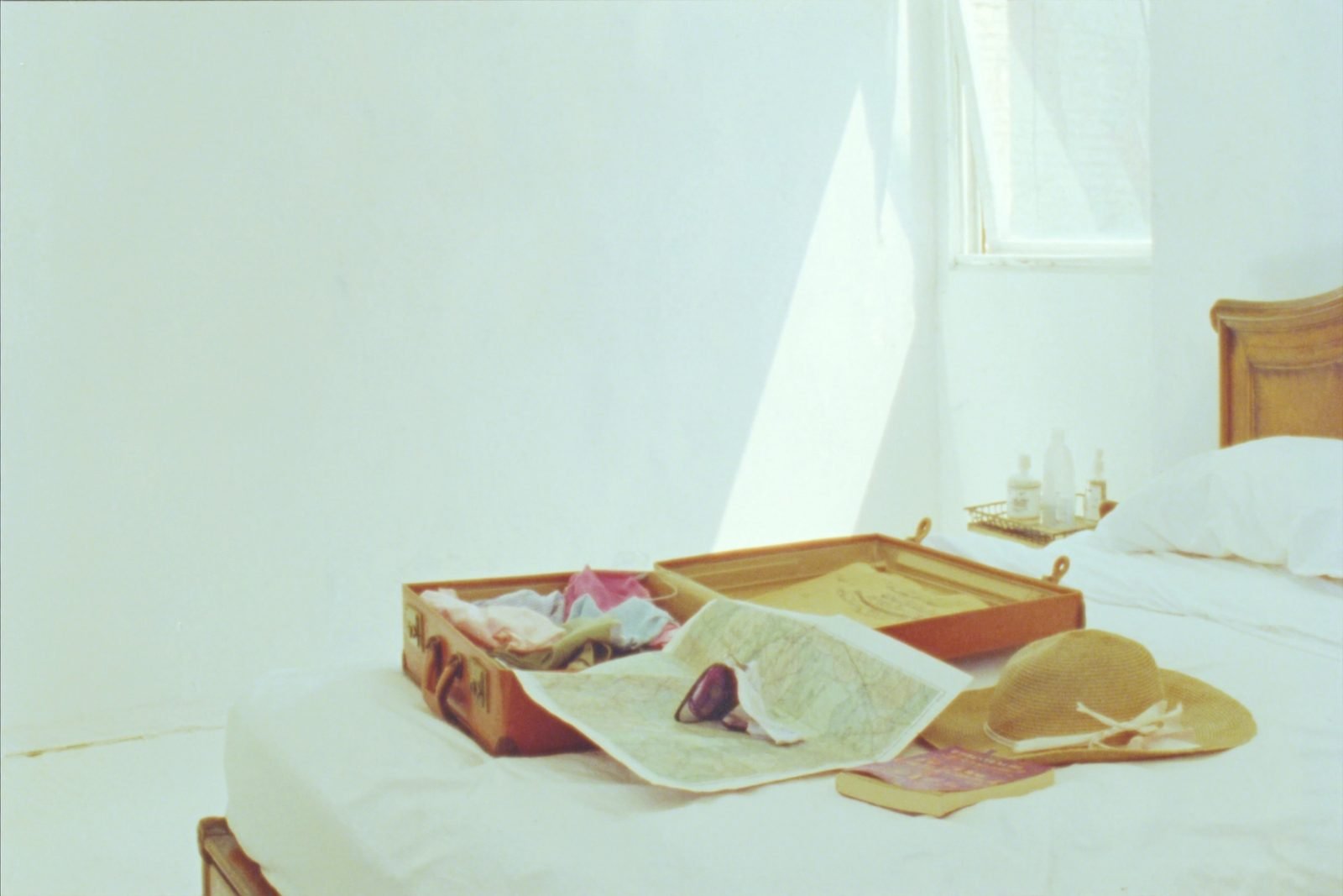 An open suitcase half packed on a white linen bed with a straw hat and a worn map