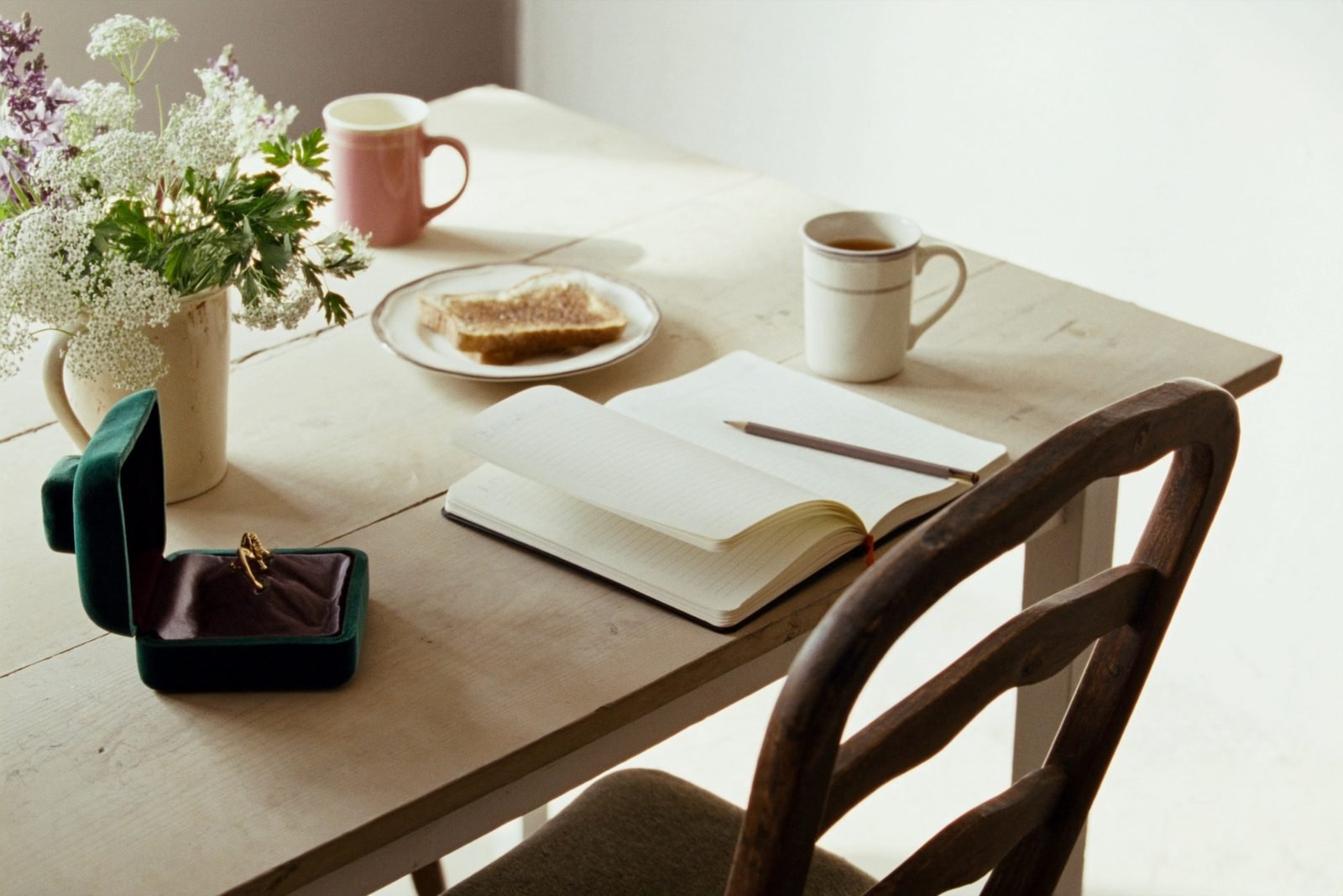 A farmhouse kitchen table in the morning with an open notebook, a ring box and two mugs of tea