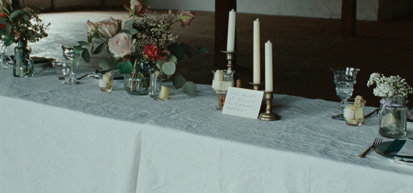 A long table laid on linen in a barn with candles, garden roses and mismatched glassware