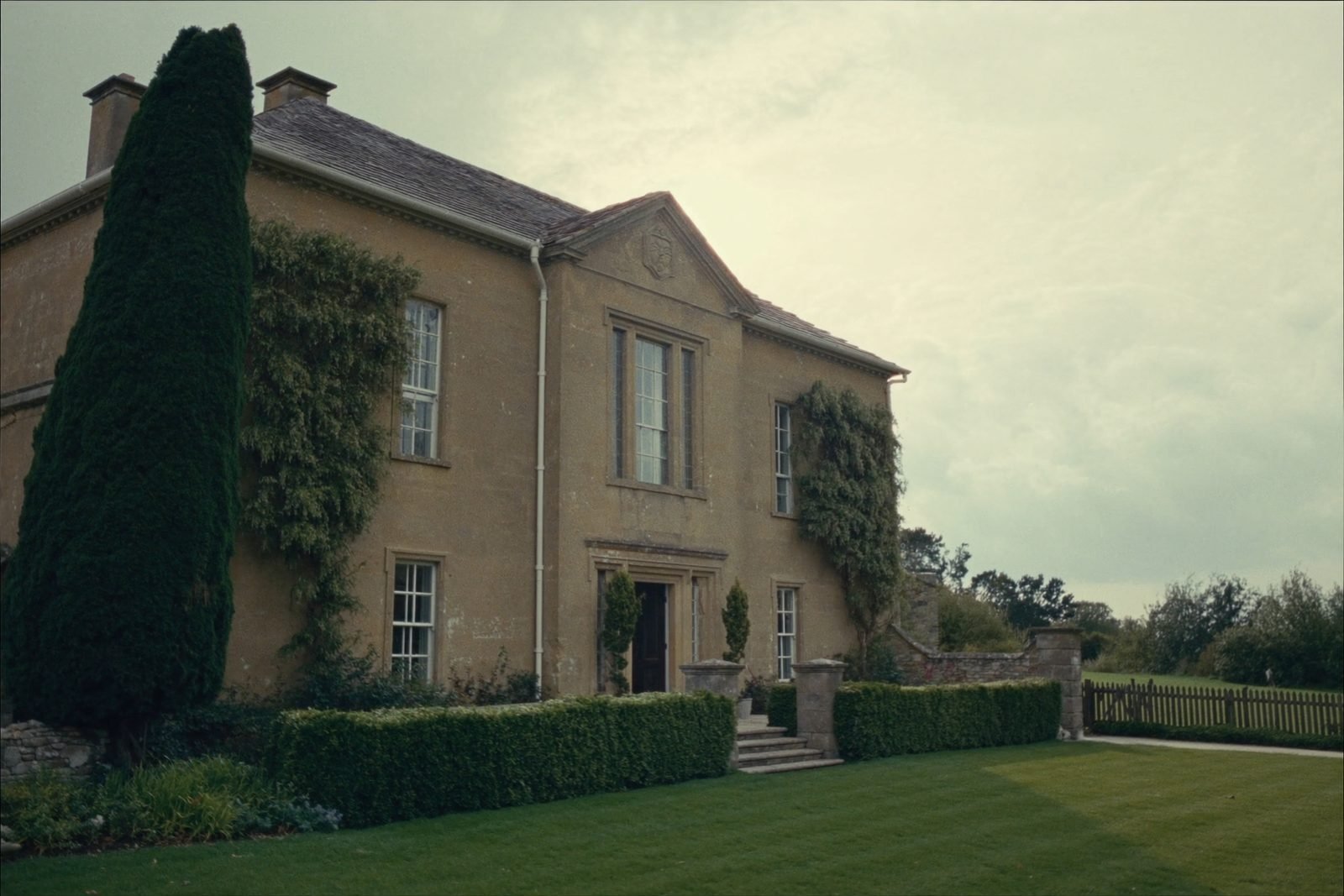 A honey coloured Cotswold stone manor house seen across a lawn with clipped yew